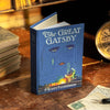 A blue book-style wallet featuring 'The Great Gatsby' by F. Scott Fitzgerald on the cover, placed on a table with a clock and other items.