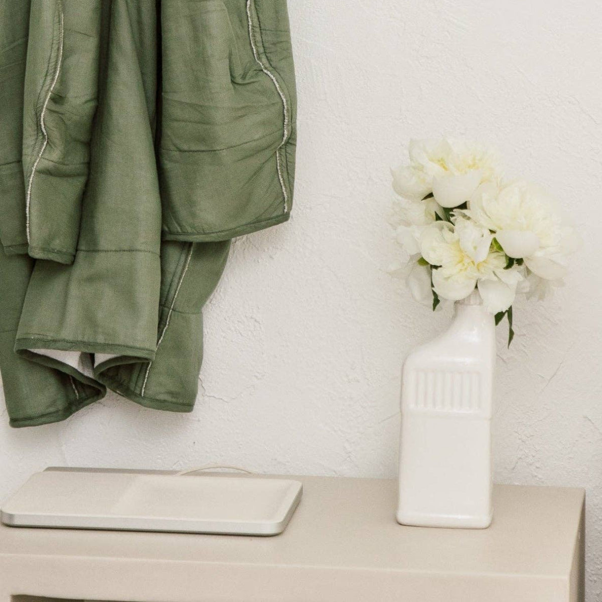 Green jacket hanging on a white wall with a vase of flowers on a table below.
