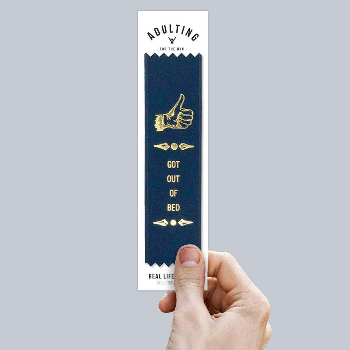 A hand holding a blue and gold satin ribbon bookmark with stamped gold foil text that reads 'GOT OUT OF BED'.