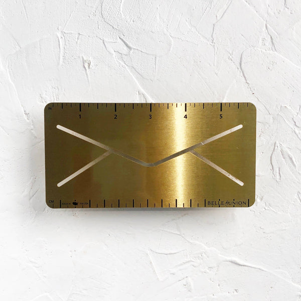 A brass envelope-shaped ruler measuring 6 inches long, with both imperial and metric markings, placed on a white background.