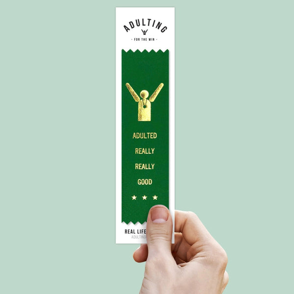 Hand holding a green award ribbon with gold text that reads "Adulted Really Really Good"