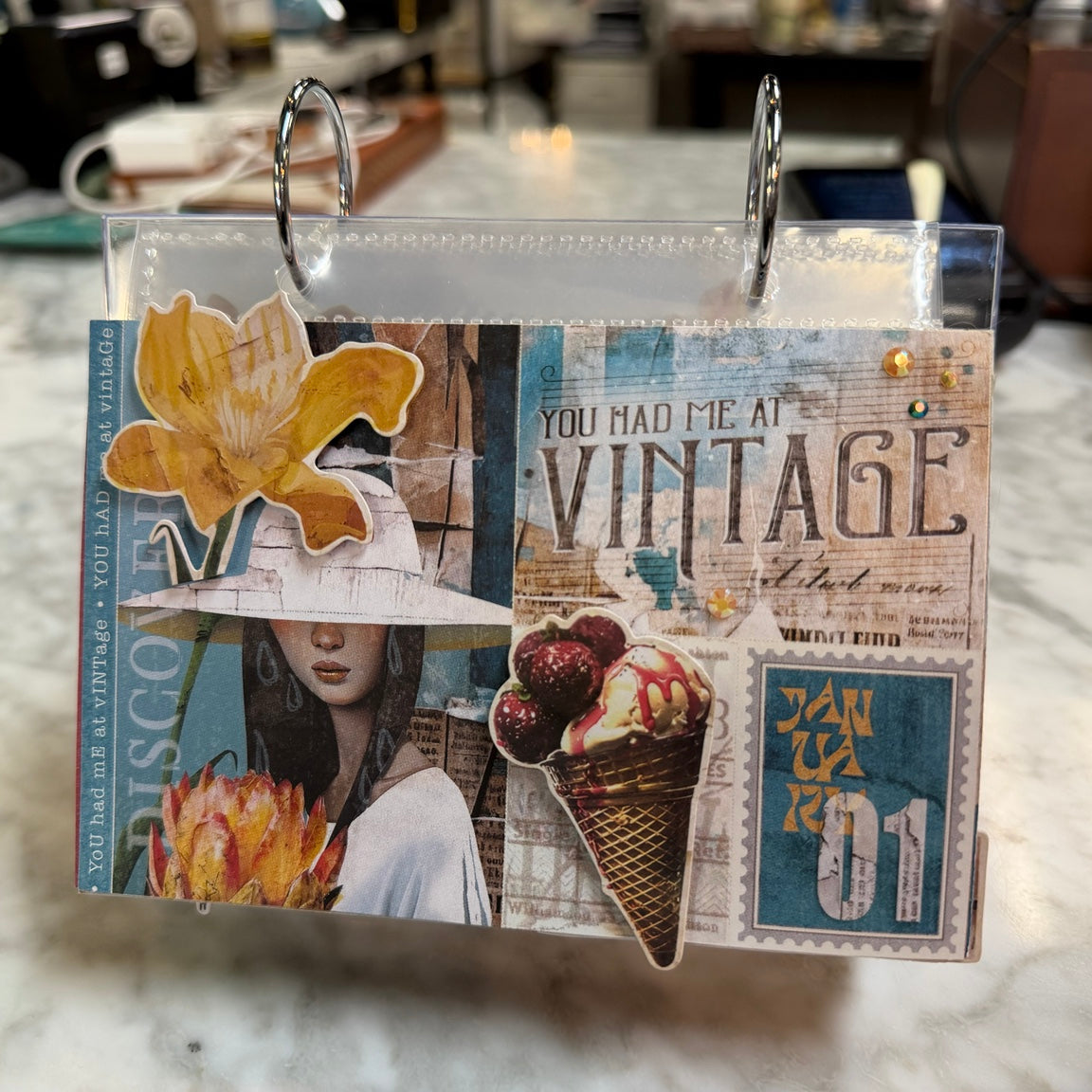 A handmade vintage-style scrapbook album or binder page with collage elements, including a yellow flower, a woman in a hat, an ice cream cone, and text like 'YOU HAD ME AT VINTAGE' and 'JAN 01', photographed hanging on a marble countertop.