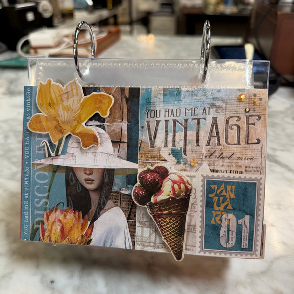 A handmade vintage-style scrapbook album or binder page with collage elements, including a yellow flower, a woman in a hat, an ice cream cone, and text like 'YOU HAD ME AT VINTAGE' and 'JAN 01', photographed hanging on a marble countertop.