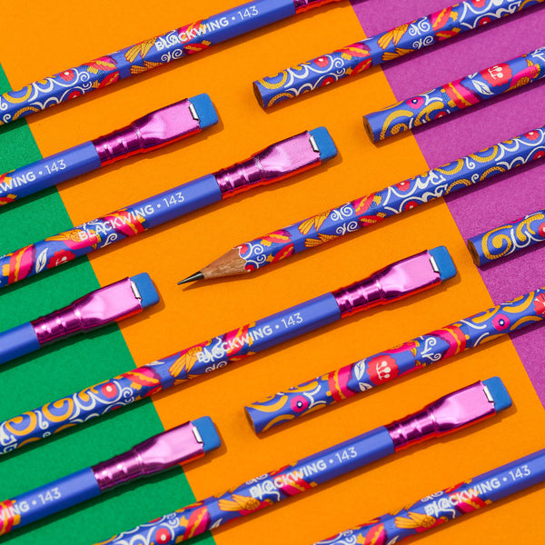 A set of colorful mechanical pencils arranged diagonally on a vibrant, geometric background of orange, green, and purple blocks. The pencils feature blue bodies with a busy, multicolored floral and swirl pattern, metallic pink erasers, and white text reading 'BLACKWING 2143'.