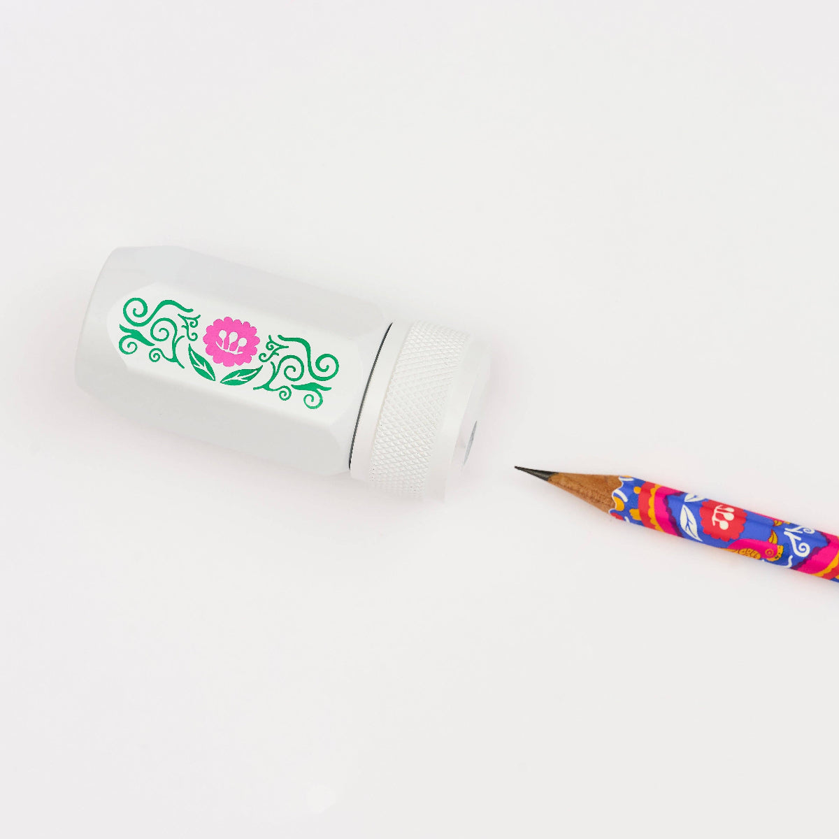 A white pencil sharpener with a green and pink floral design, shown next to a colorful patterned pencil on a clean white background.