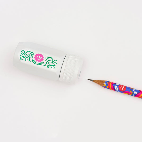 A white pencil sharpener with a green and pink floral design, shown next to a colorful patterned pencil on a clean white background.