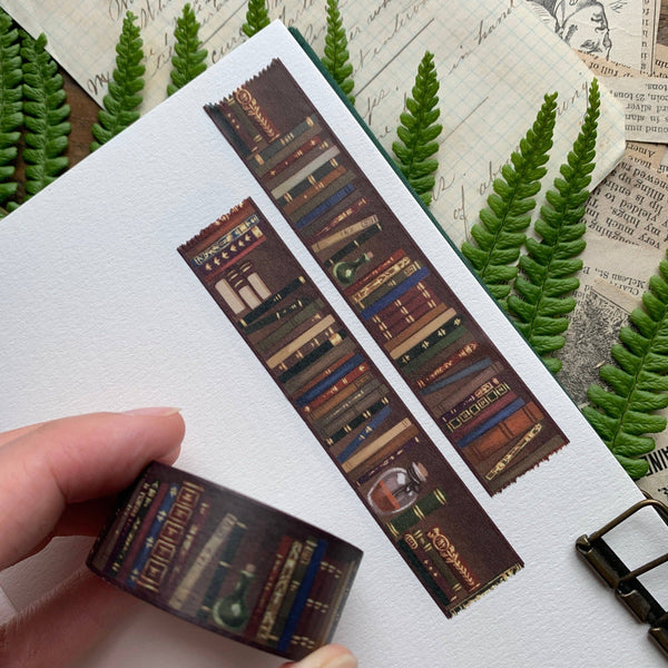Decorative book-themed washi tape with a hand holding it, set against a textured background with greenery.
