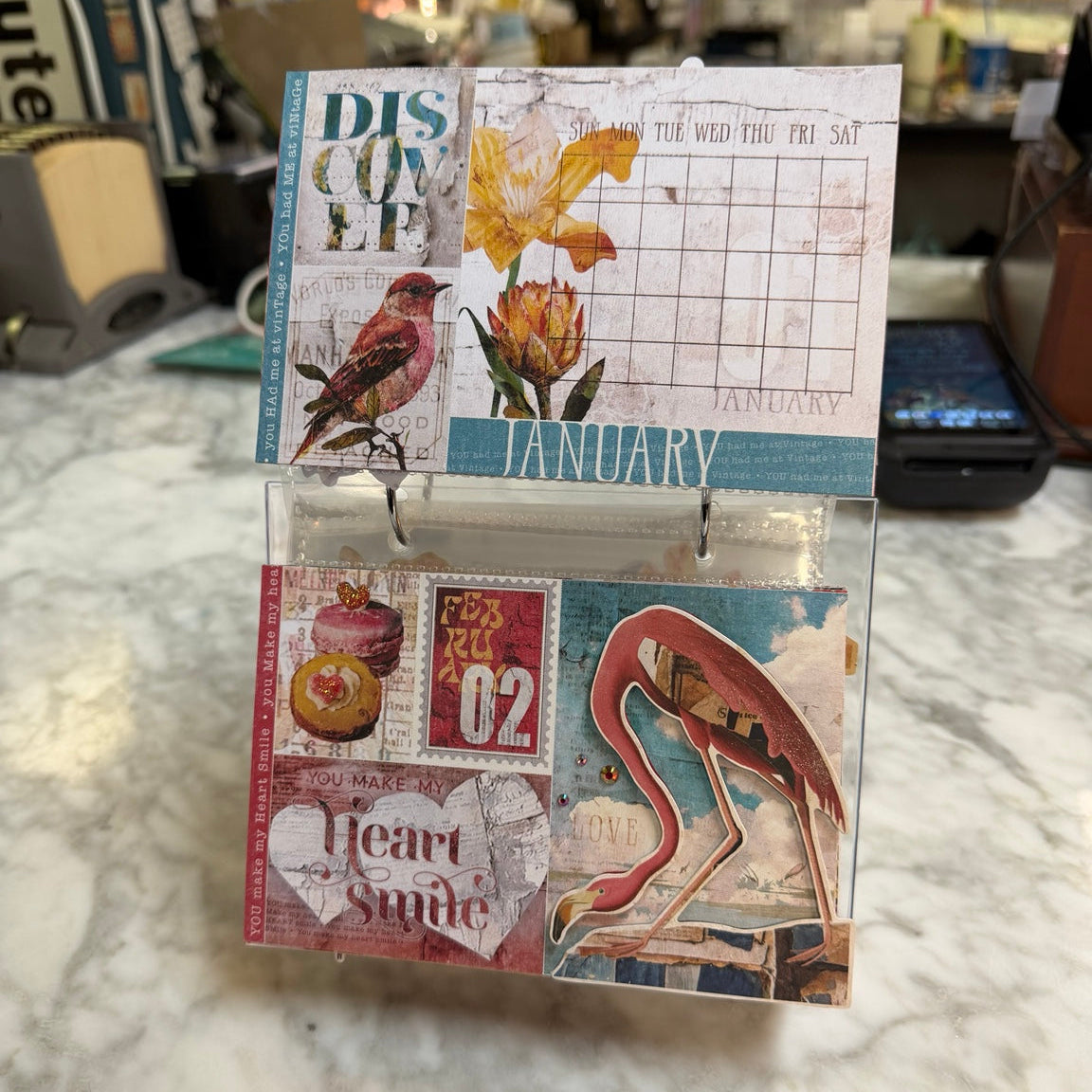 A small decorative scrapbook/card collage kit displayed open on a marble countertop in a real store environment. The kit includes vintage-style printed panels with a bird, flowers, a calendar grid, stamps, and a flamingo illustration.