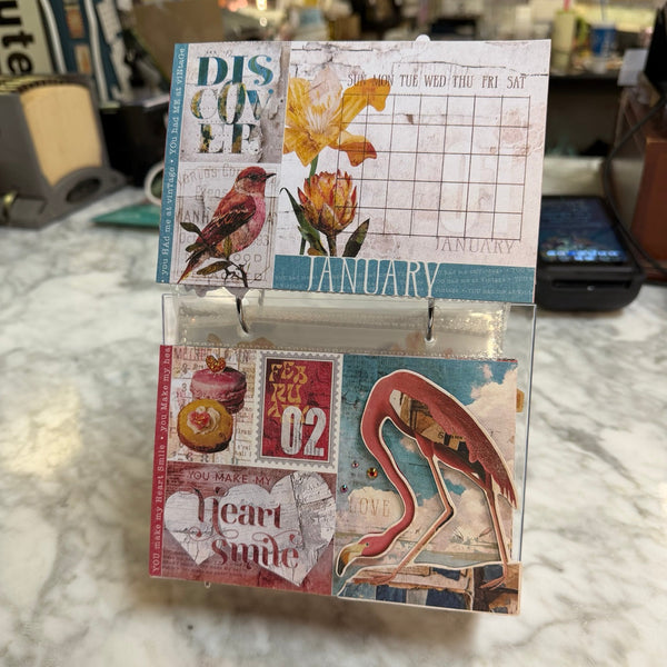 A small decorative scrapbook/card collage kit displayed open on a marble countertop in a real store environment. The kit includes vintage-style printed panels with a bird, flowers, a calendar grid, stamps, and a flamingo illustration.