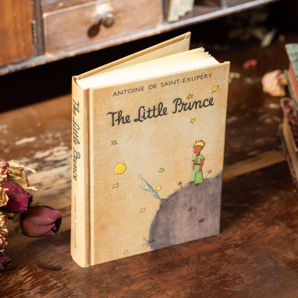 Book titled 'The Little Prince' by Antoine de Saint-Exupéry on a wooden surface with flowers and a vintage cabinet in the background.