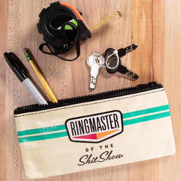 A beige canvas zippered pouch with green stripes and a 'RINGMASTER OF THE Shit Show' logo, styled flat-lay on a wooden surface with keys, a tape measure, and pens.