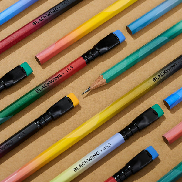 A set of colorful mechanical pencils arranged diagonally on a textured kraft paper background. The pencils feature vibrant gradient bodies (red, orange, yellow, green, blue, pink) with black ferrules, black erasers, and colored rubber tips in blue, green, yellow, and red.