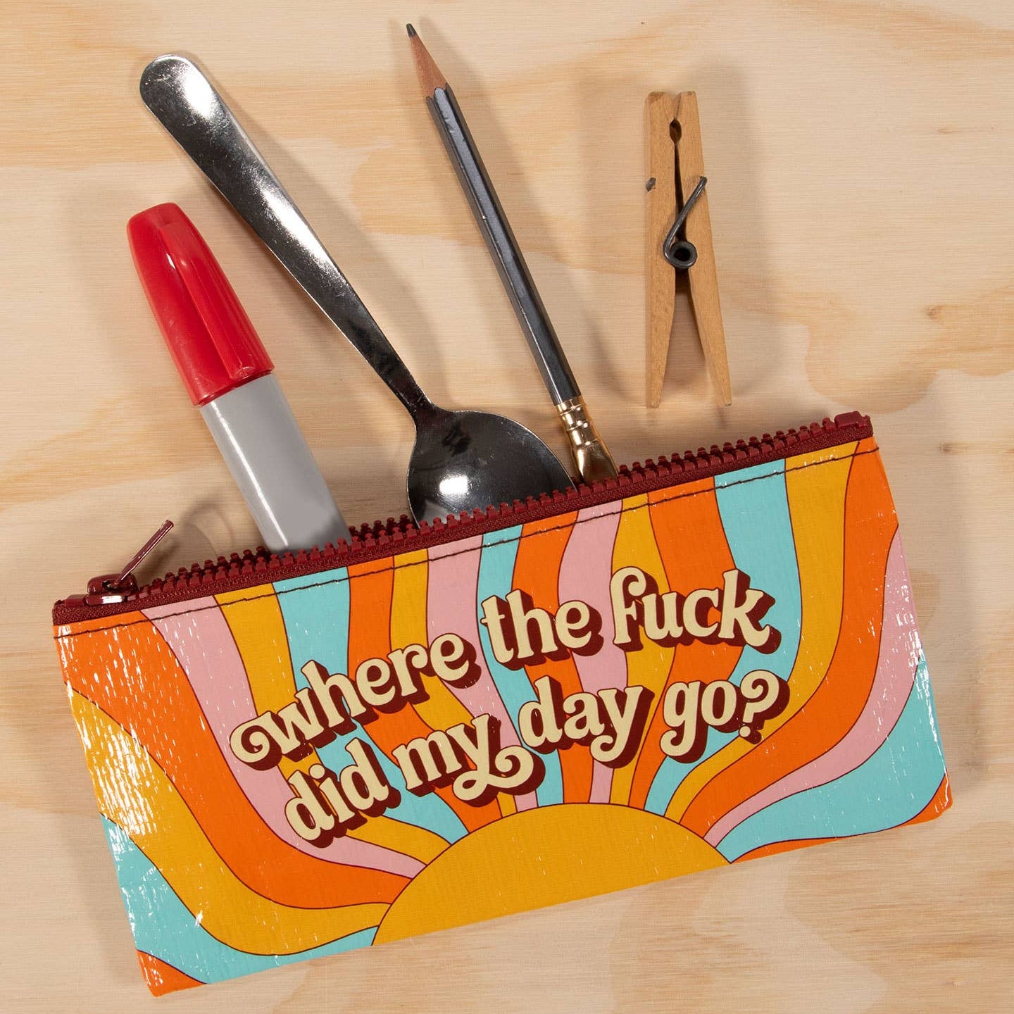 A colorful zippered pouch with a retro sunburst design and the text 'where the fuck did my day go?' lying on a light wood surface, with pens, a spoon, a pencil, and a wooden clothespin peeking out from the top.