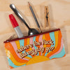 A colorful zippered pouch with a retro sunburst design and the text 'where the fuck did my day go?' lying on a light wood surface, with pens, a spoon, a pencil, and a wooden clothespin peeking out from the top.
