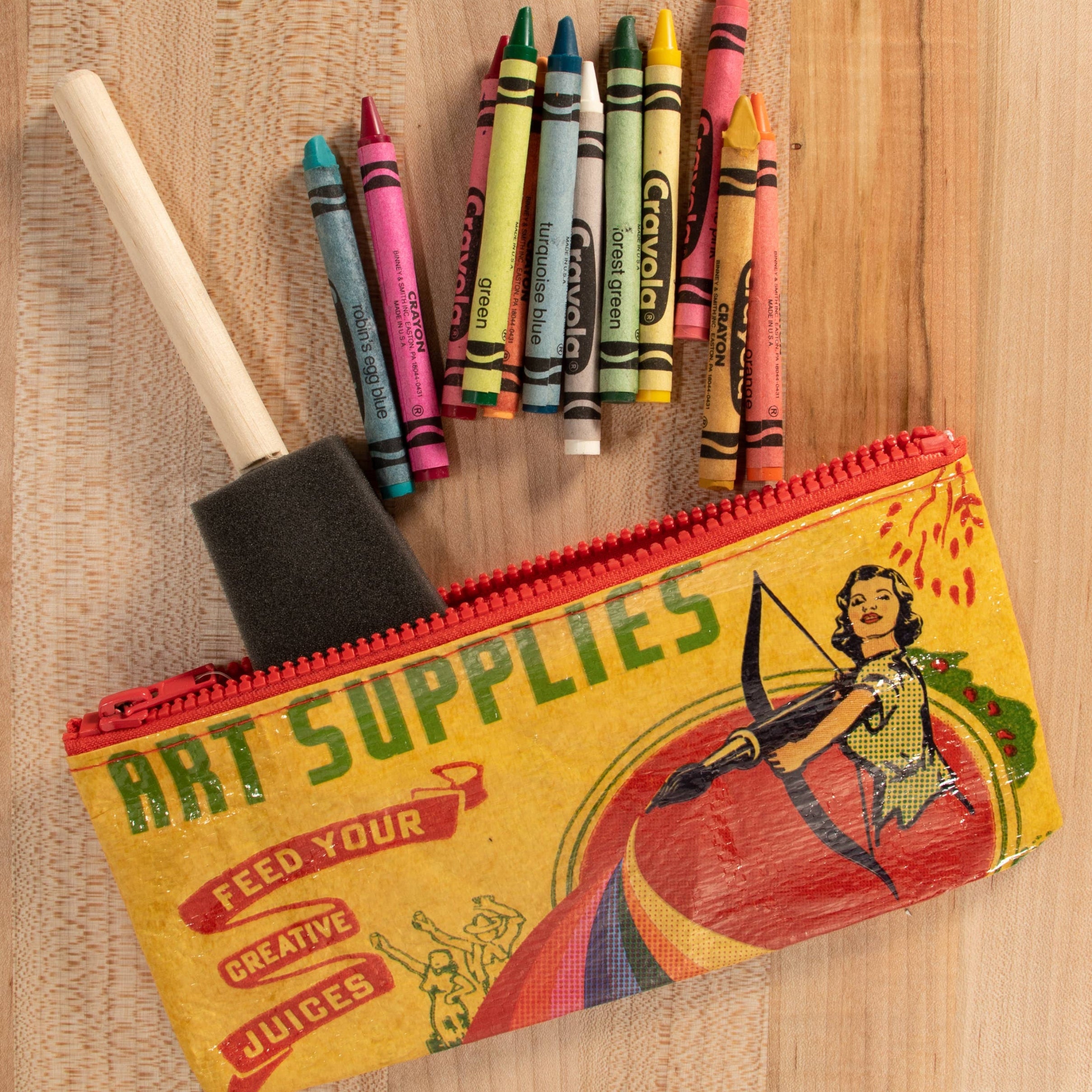A vintage-style illustrated pencil case labeled 'ART SUPPLIES' lies on a light wood surface, with crayons and a paintbrush placed on top of it.