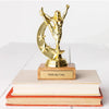 A trophy with a figurine standing with arms raised on top of a wooden base, placed on top of two books. The trophy has a brass-look plaque with the phrase 'FUCK ALL Y'ALL' engraved on it.