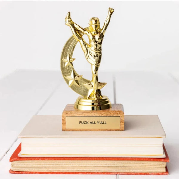 A trophy with a figurine standing with arms raised on top of a wooden base, placed on top of two books. The trophy has a brass-look plaque with the phrase 'FUCK ALL Y'ALL' engraved on it.